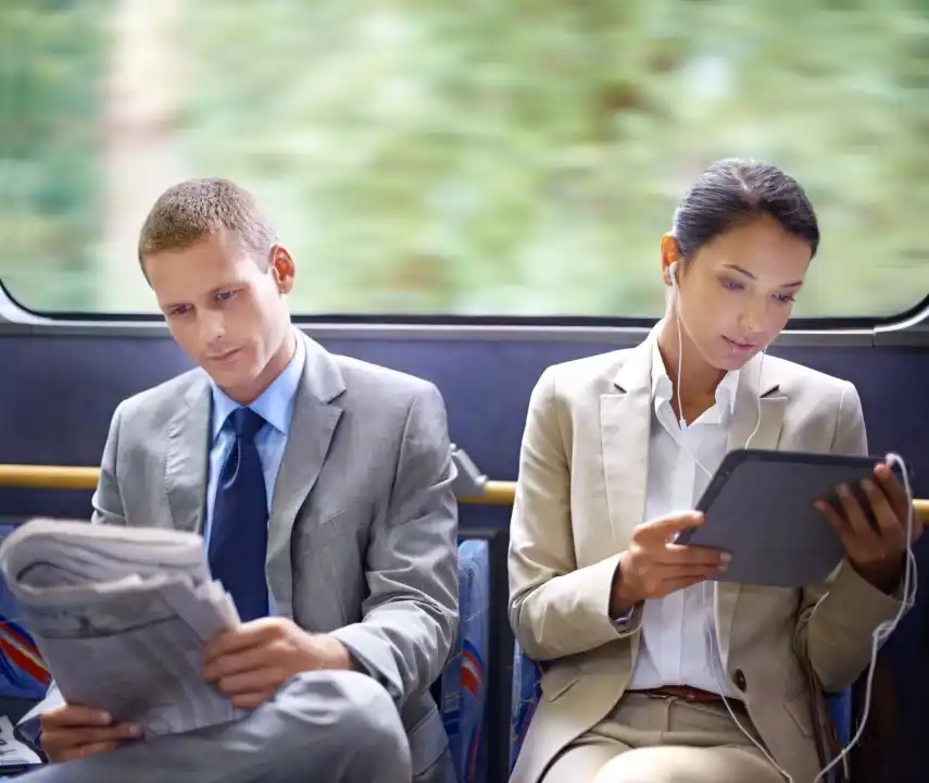 Two professionals commuting on a bus—man reading a newspaper, woman using a tablet with earphones—illustrating modern digital media consumption.