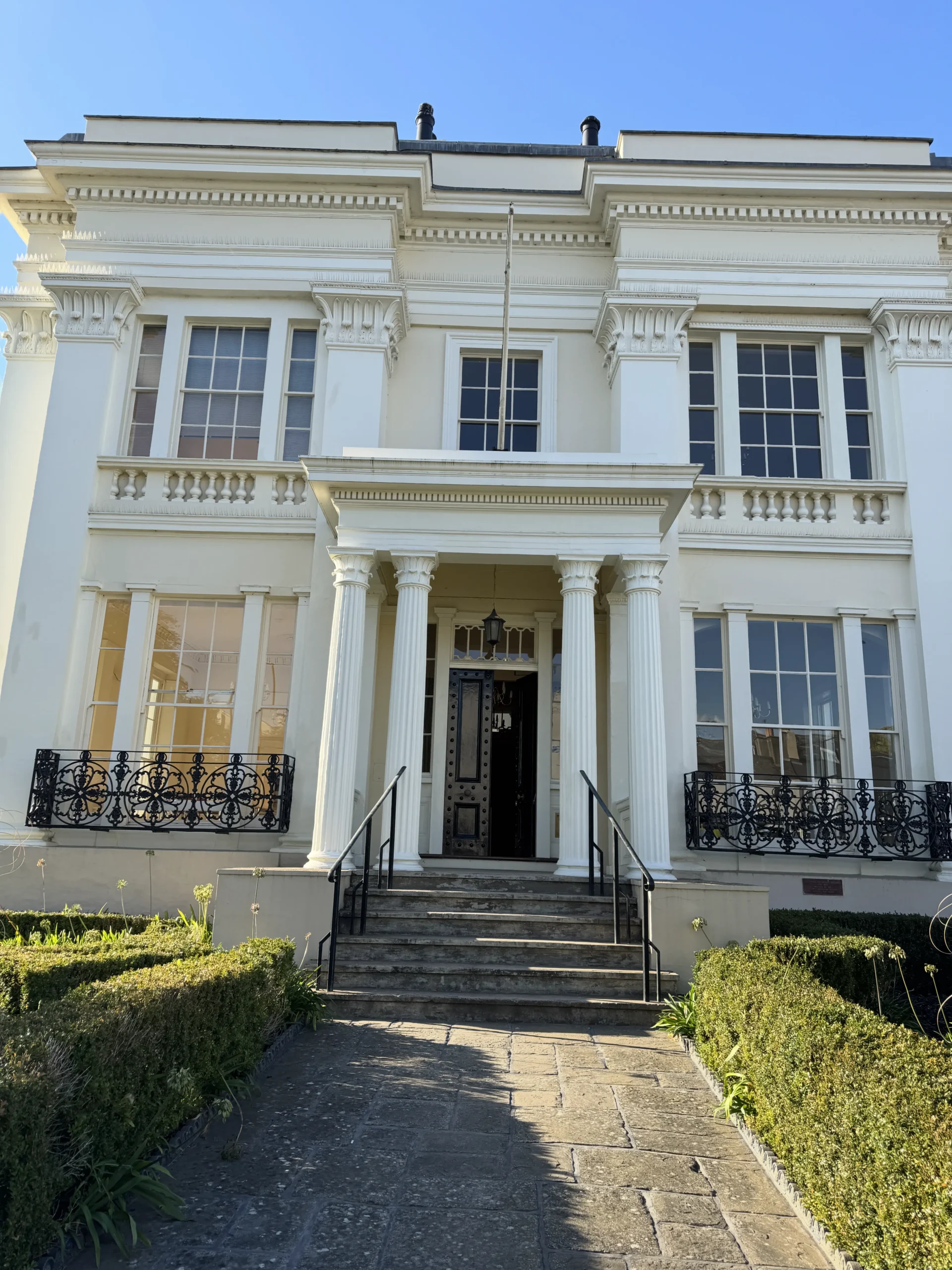 front view of Lauriston House, a white neoclassical building with tall columns, black iron railings, and manicured hedges, now home to our new offices on Montpellier Street, Cheltenham