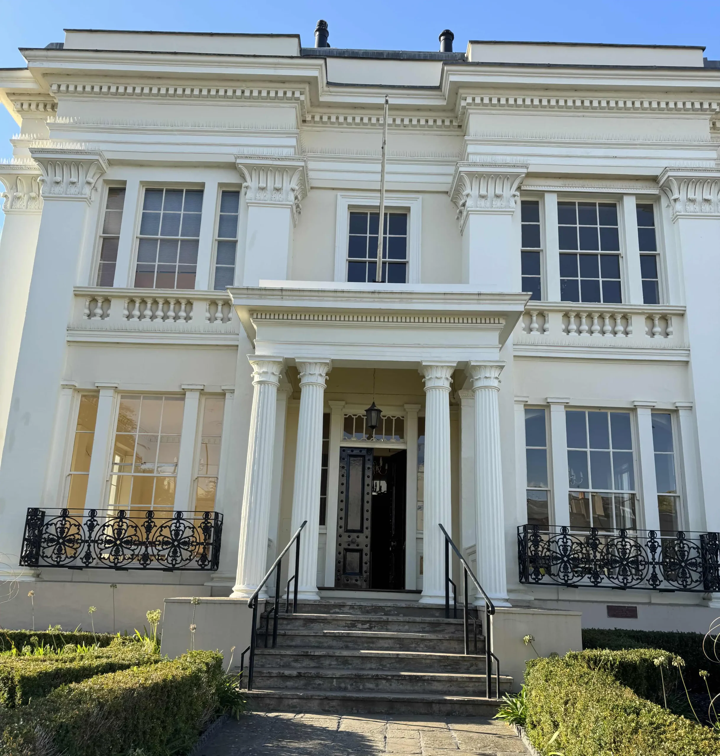 front view of Lauriston House, a white neoclassical building with tall columns, black iron railings, and manicured hedges, now home to our new offices on Montpellier Street, Cheltenham